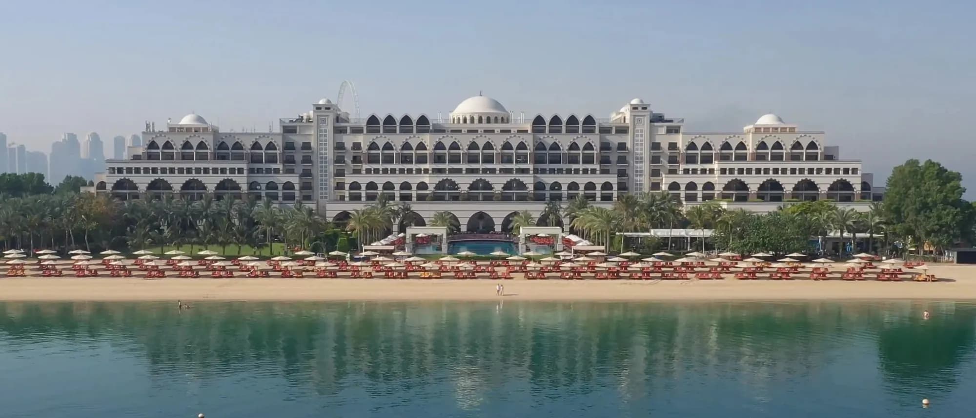 A palace-style hotel with white domes and arches behind a beach and calm water