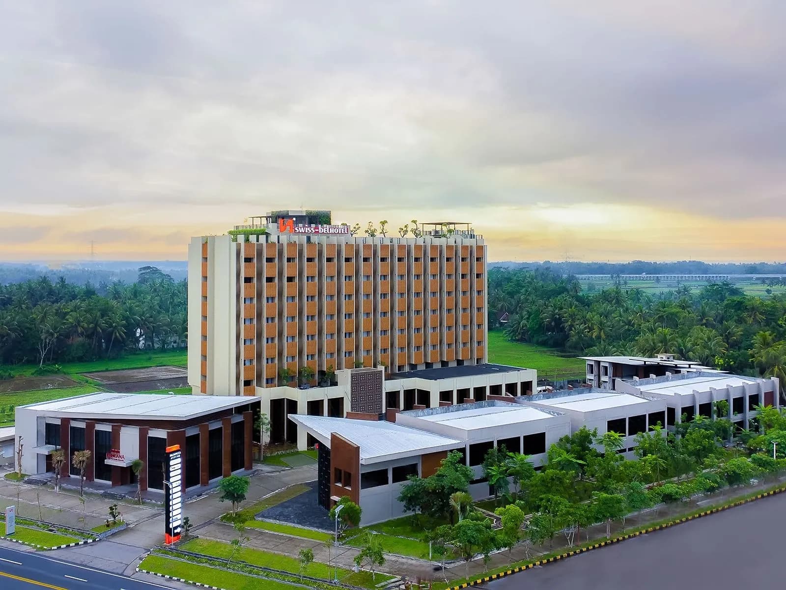 A tall hotel tower with low wings in front, set among trees and green fields at sunset